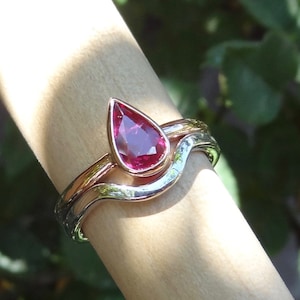 May include: A pear-shaped ruby gemstone set in a rose gold ring, with a wavy silver band underneath.