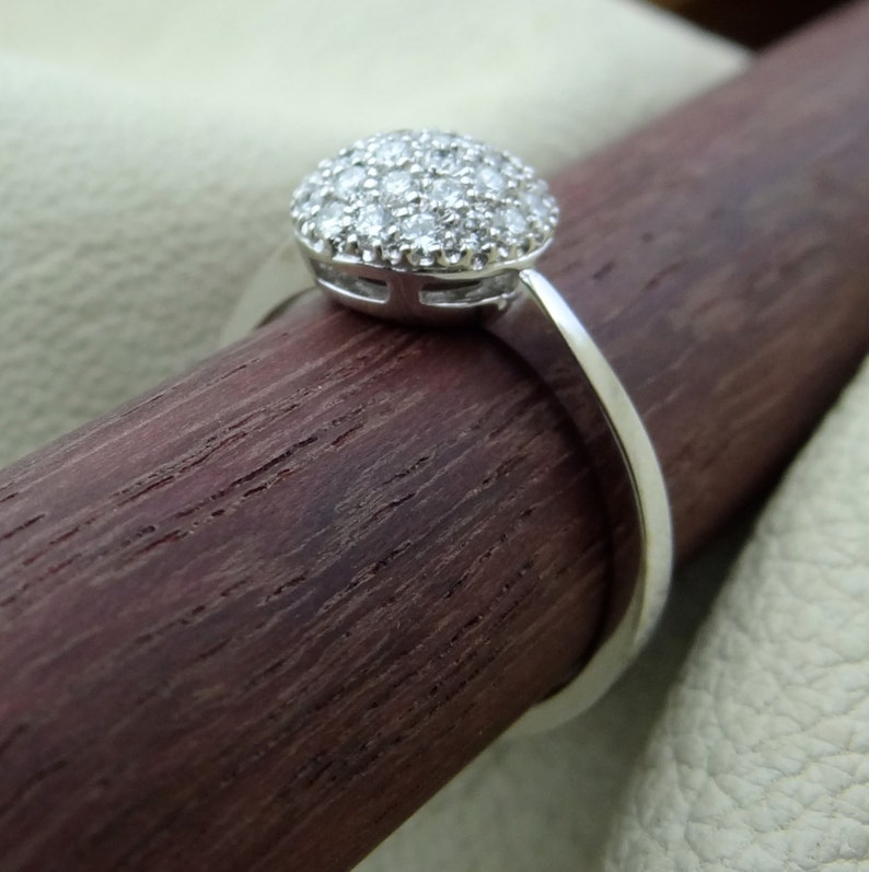 May include: A silver ring with a round, pave-set diamond center stone. The ring is on a wooden surface.
