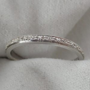 May include: A silver ring with an intricate floral pattern. The ring has a delicate design with small, detailed flowers and leaves. The band is smooth and polished, with a subtle, textured border. The ring is likely made of sterling silver or a similar metal.