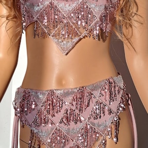 K&ouml;nnte beinhalten: Ein zweiteiliges Outfit mit einem rosa paillettenbesetzten Oberteil und einem passenden Unterteil. Das Oberteil ist ein Bandeau-Stil mit Fransen-Details. Das Unterteil ist ein hoch taillierter Stil mit Fransen-Details und B&auml;ndern an den Seiten.