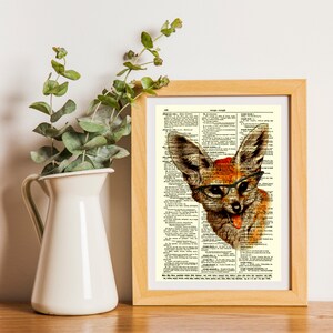 May include: A framed print featuring a whimsical illustration of a fox wearing glasses and a red hat, set against a vintage dictionary page. The artwork is in a wooden frame, with a white pitcher and green foliage in the background.