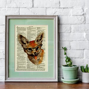 May include: Framed artwork featuring a whimsical fox illustration wearing glasses, set against a vintage dictionary page. The fox is depicted with orange and brown fur, a pink tongue, and black-rimmed glasses. The artwork is framed in a light green mat and a gray frame.
