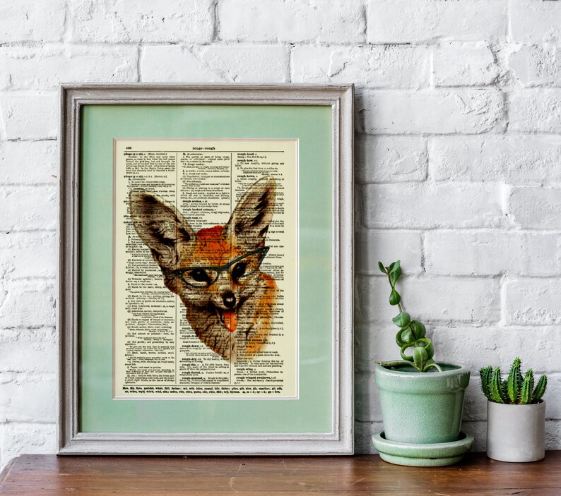 May include: Framed artwork featuring a whimsical fox illustration wearing glasses, set against a vintage dictionary page. The fox is depicted with orange and brown fur, a pink tongue, and black-rimmed glasses. The artwork is framed in a light green mat and a gray frame.