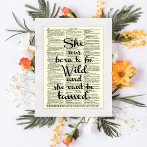 May include: A framed print with the quote "She was born to be Wild and she can't be tamed" in black script. The print is set against a vintage dictionary page, surrounded by white flowers, orange roses, and greenery.