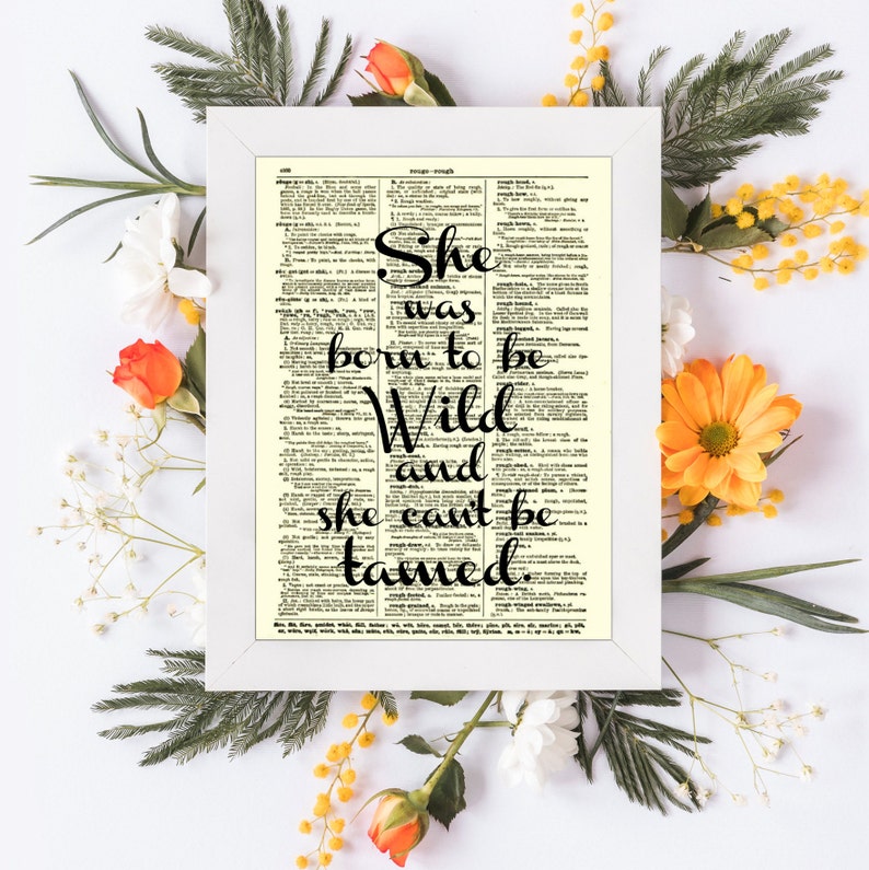 May include: A framed print with the quote "She was born to be Wild and she can't be tamed" in black script. The print is set against a vintage dictionary page, surrounded by white flowers, orange roses, and greenery.