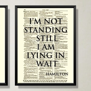 以下が含まれることがあります： 黒いフレームのアートプリント。テキスト「I'M NOT STANDING STILL I AM LYING IN WAIT HAMILTON」がヴィンテージ辞書ページに表示されています。