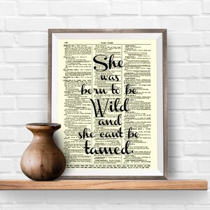 May include: A framed print with the words "She was born to be Wild and she can't be tamed" in black script. The print is set against a dictionary page background. A wooden vase sits on a wooden shelf below the print.