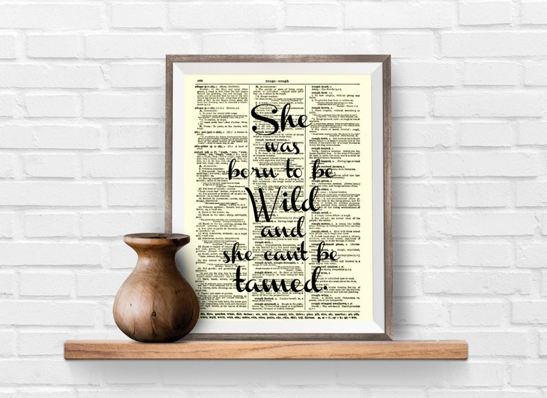 May include: A framed print with the words "She was born to be Wild and she can't be tamed" in black script. The print is set against a dictionary page background. A wooden vase sits on a wooden shelf below the print.
