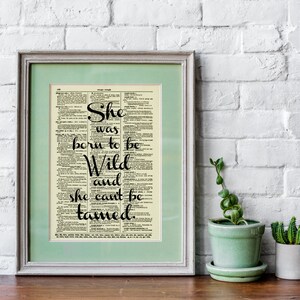 May include: Framed print featuring the quote "She was born to be Wild and she can't be tamed" in elegant black script. The text is set against a vintage dictionary page background, framed in a light green mat and a silver frame. A decorative piece.