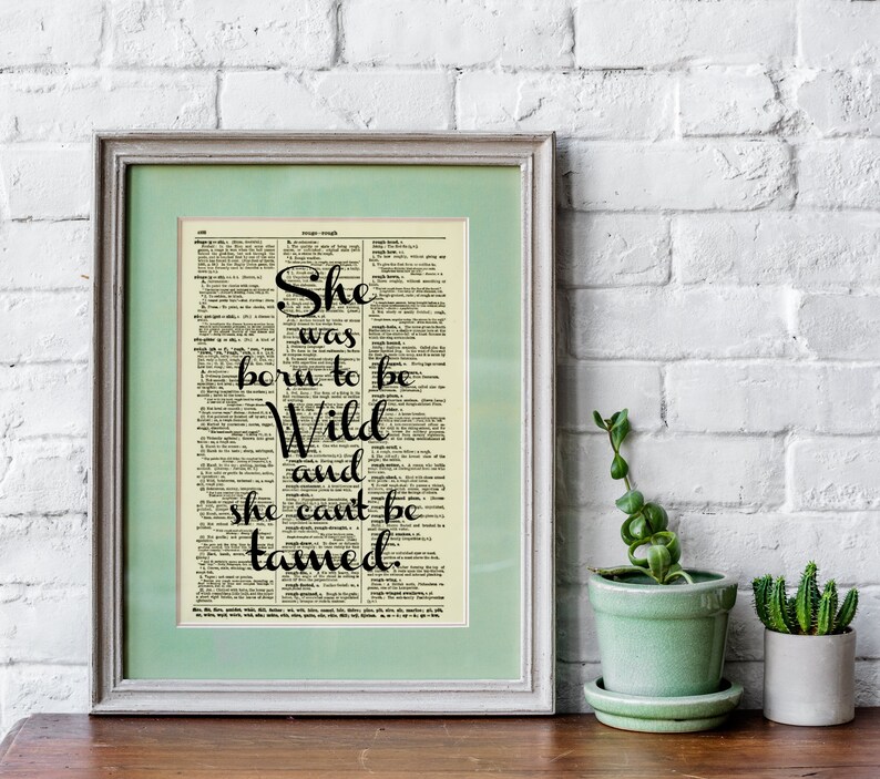 May include: Framed print featuring the quote "She was born to be Wild and she can't be tamed" in elegant black script. The text is set against a vintage dictionary page background, framed in a light green mat and a silver frame. A decorative piece.