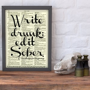 以下が含まれることがあります： ヴィンテージ辞書ページを背景にした額入りのプリント。黒いテキストは、アーネスト・ヘミングウェイによる「Write drunk; edit sober」と書かれています。フレームは濃いグレーです。背景は白いレンガの壁です。