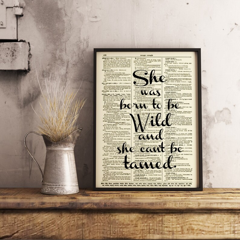 May include: A framed print with the quote "She was born to be Wild and she can't be tamed" in black script. The background is a vintage dictionary page. A metal pitcher with dried grass sits on a wooden shelf.