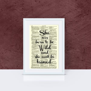 May include: A framed print featuring the quote "She was born to be Wild and she can't be tamed." The text is in black script over a vintage dictionary page, set in a white frame. The background is a deep burgundy.