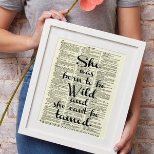 May include: White framed print featuring the quote "She was born to be Wild and she can't be tamed" in black script. The background is a vintage dictionary page. A pink flower is held near the frame.