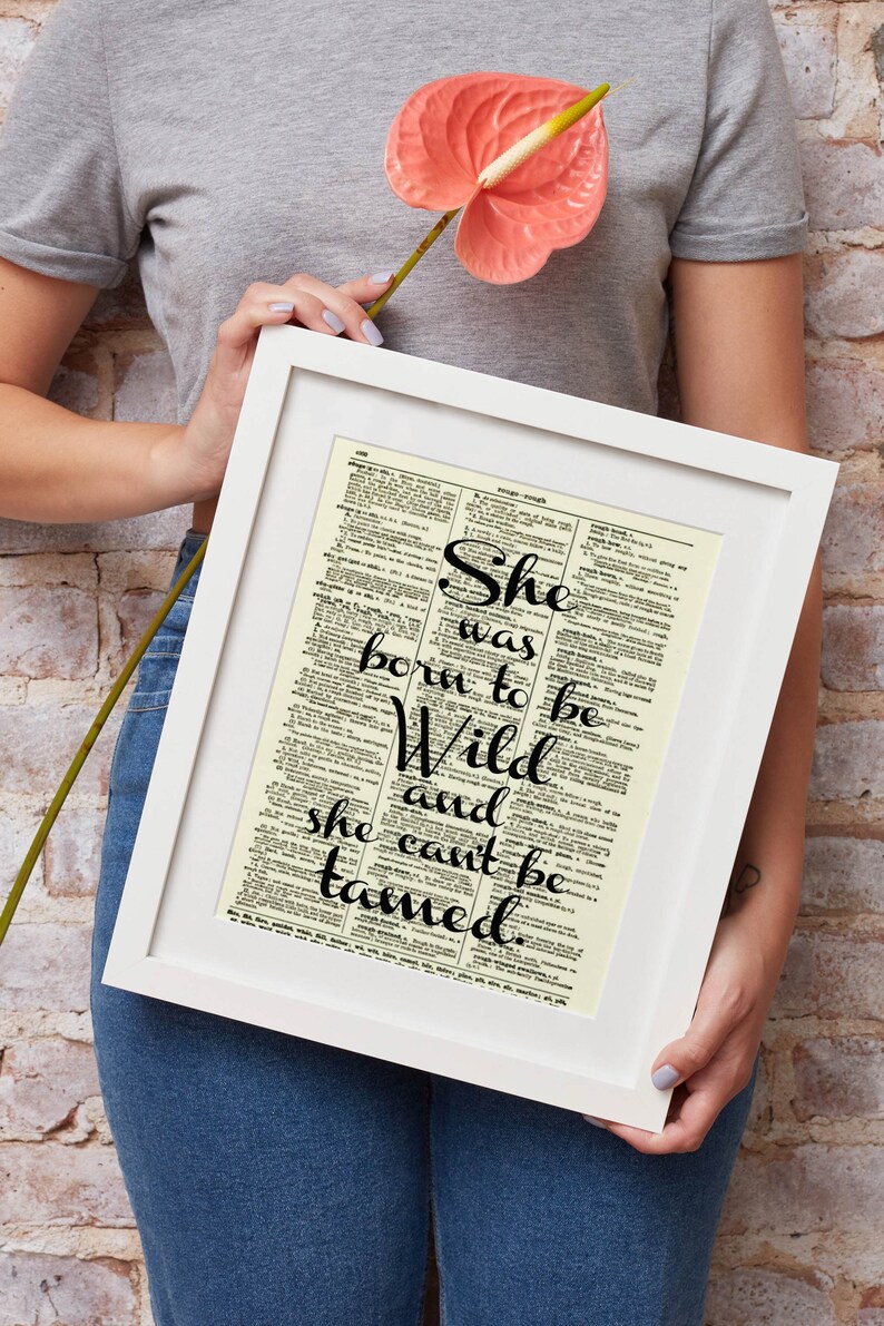 May include: White framed print featuring the quote "She was born to be Wild and she can't be tamed" in black script. The background is a vintage dictionary page. A pink flower is held near the frame.
