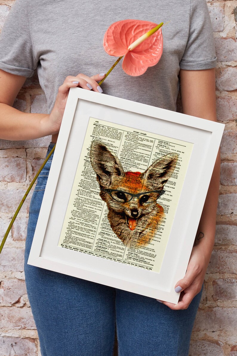 May include: A framed art print featuring a whimsical illustration of a fox wearing glasses and sticking out its tongue. The artwork is set against a vintage dictionary page background, with a white frame. The fox is depicted in shades of brown and orange.
