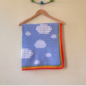 Peut inclure: Une couverture bébé en crochet bleu clair avec des nuages blancs et une bordure arc-en-ciel. La couverture est suspendue à un cintre en bois.