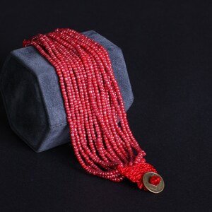 May include: A multi-strand red beaded bracelet with a gold-colored clasp. The bracelet is draped over a gray, geometric display block against a black background. The beads are small and uniform in size.