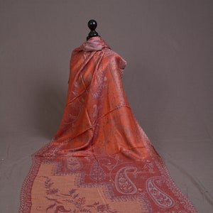 May include: An orange paisley patterned scarf draped over a mannequin. The scarf features a detailed design with shades of orange, red, and purple. The scarf has a fringed edge and is made of a soft, flowing fabric.