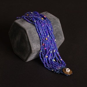 May include: A multi-strand bracelet featuring strands of small, iridescent blue beads. The bracelet has a decorative clasp with a clear stone and a gold-colored metal accent. The beads are interspersed with small, colorful beads.