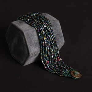 May include: A multi-strand beaded bracelet with iridescent beads in shades of green, blue, and purple. The bracelet is secured with a green clasp and a decorative gold-colored button. The jewelry is displayed on a gray, geometric block against a black background.