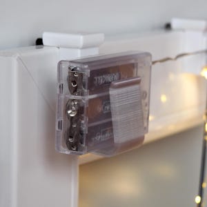 Könnte beinhalten: Eine transparente Batteriebox mit drei Duracell AA-Batterien, befestigt an einer weißen Oberfläche. Die Box ist an einer weißen Struktur befestigt, im Hintergrund ist eine Lichterkette mit kleinen, warmtonigen Lichtern zu sehen.