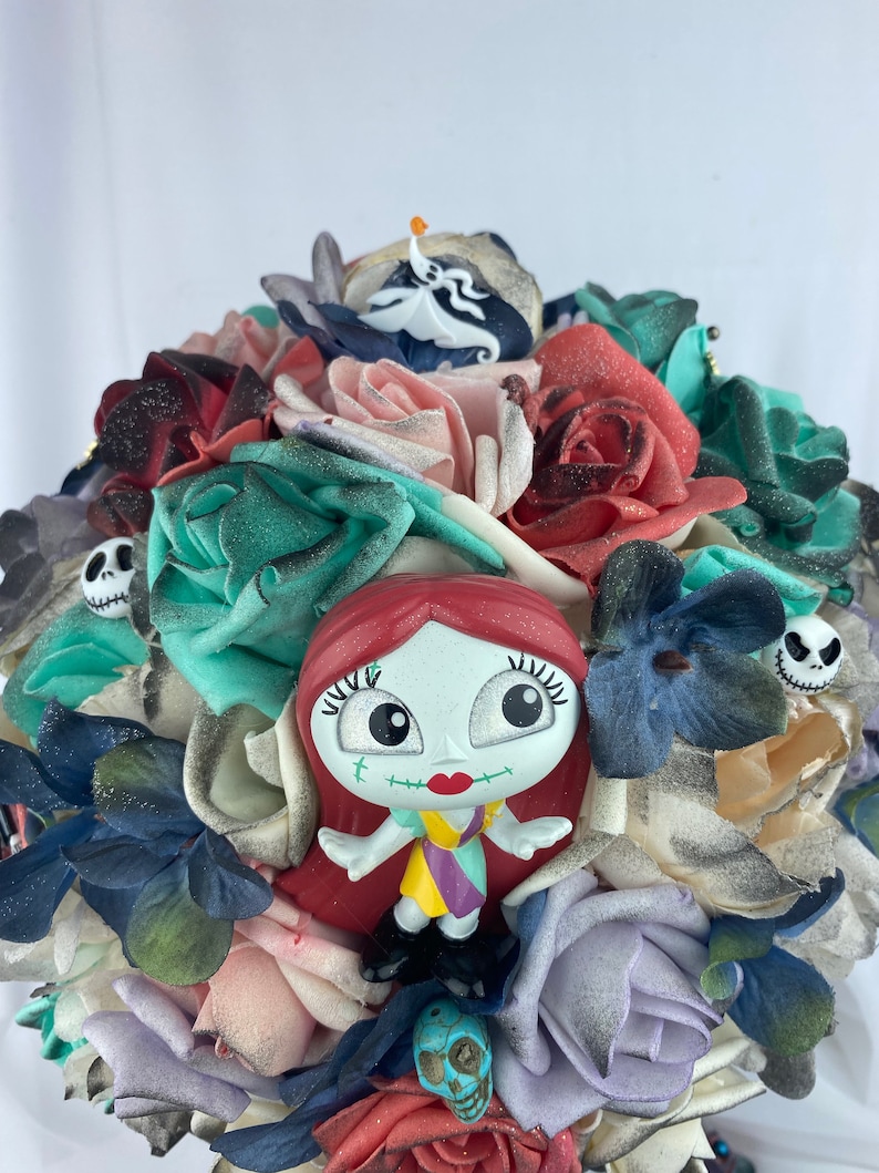 May include: A bouquet of artificial flowers in shades of red, pink, blue, and green. The bouquet features a small plastic figurine of Sally from the Nightmare Before Christmas, a skull, and a ghost.