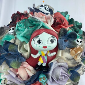 May include: A bouquet of artificial flowers in shades of red, pink, blue, and green. The bouquet features a small plastic figurine of Sally from the Nightmare Before Christmas, surrounded by skulls and other Halloween-themed decorations.