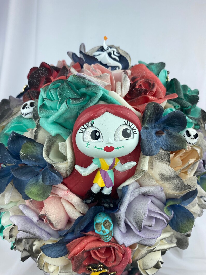 May include: A bouquet of artificial flowers in shades of red, pink, blue, and green. The bouquet features a small plastic figurine of Sally from the Nightmare Before Christmas, surrounded by skulls and other Halloween-themed decorations.