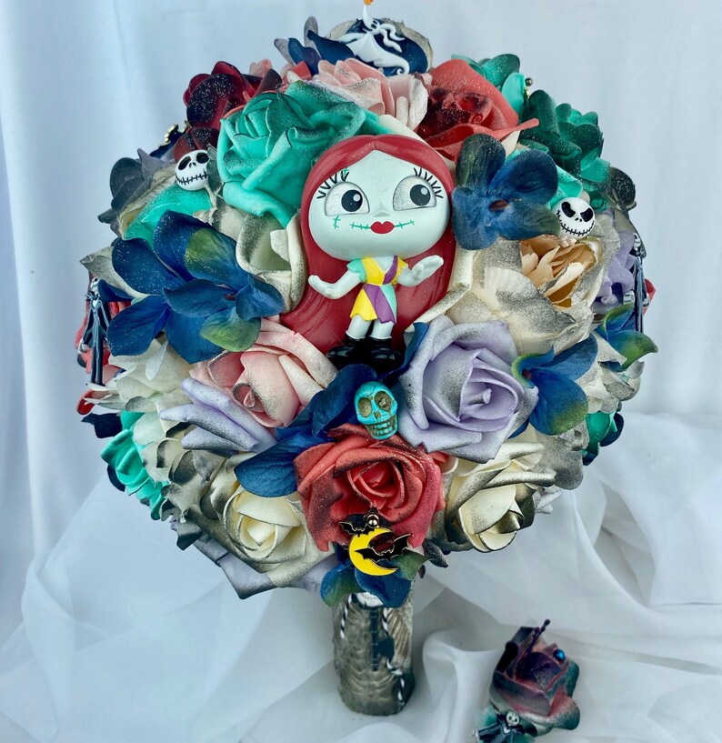 May include: A colorful bouquet of artificial flowers with a small plastic doll of Sally from the Nightmare Before Christmas movie. The bouquet is decorated with various Halloween-themed charms, including skulls and bats.