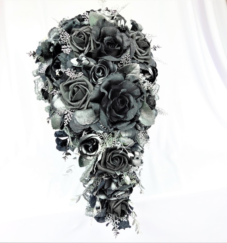 May include: A cascading bridal bouquet made of black roses with silver glitter accents. The bouquet is arranged in a teardrop shape and features a variety of black roses, some with a more velvety texture and others with a more satin finish.