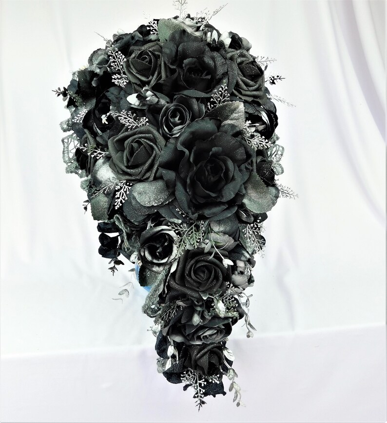May include: A cascading bridal bouquet with black roses and silver accents. The bouquet is made of artificial flowers and is perfect for a gothic wedding.