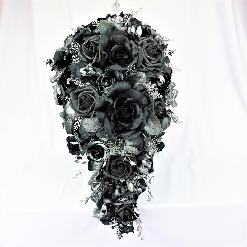 May include: A cascading bridal bouquet made of black roses and silver foliage. The bouquet is arranged in a teardrop shape and features a variety of black roses, some with glitter accents.