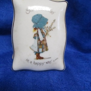 Peut inclure: Un vase rectangulaire en céramique avec une finition blanche et un bord fin foncé. L'avant présente un graphique d'une personne portant un bonnet bleu et des vêtements en patchwork tenant des fleurs, avec le texte "Start each day in a happy way".