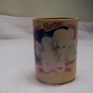 May include: A beige ceramic cup with a pink and blue design featuring a white puppy with the word "Barbie" printed on the front.