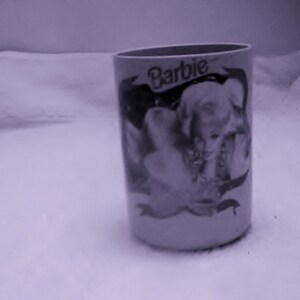 May include: A white ceramic mug with a black and white image of a woman with the word "Barbie" printed on it.