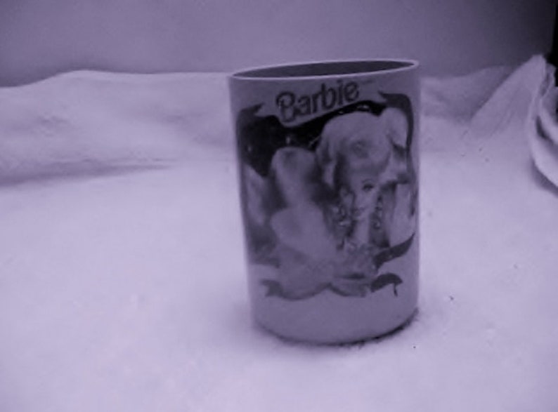 May include: A white ceramic mug with a black and white image of a woman with the word "Barbie" printed on it.