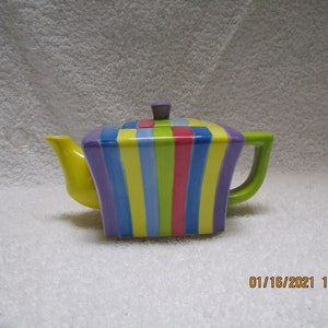 May include: A colourful striped teapot with a square body and a yellow spout. The teapot is decorated with vertical stripes in shades of purple, blue, yellow, green, and pink.