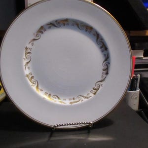 Puede incluir: Plato blanco con borde dorado y un patrón floral dorado decorativo en el borde interior. El plato se exhibe en un soporte de metal plateado. Una pieza clásica para servir o exhibir.