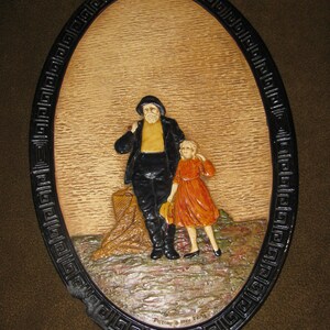 May include: A framed oval-shaped wall plaque with a wood-like background featuring a carved image of a man and a girl. The man is wearing a dark jacket, a light shirt, and a hat. The girl is wearing a red dress. The plaque has a black border with a decorative pattern.