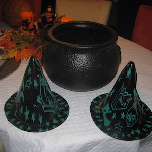 Peut inclure: Deux chapeaux de sorcière noirs avec des étoiles vertes et blanches et un motif de crâne. Les chapeaux sont sur une nappe blanche avec un chaudron noir en arrière-plan.