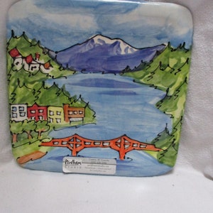 Puede incluir: Plato de cerámica pintado a mano que representa un paisaje escénico con una montaña, un lago, un puente y casas. El plato está decorado con colores brillantes y tiene un fondo de cielo azul. El texto "Priddham Studio" es visible en la esquina inferior derecha.