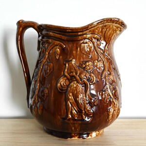 May include: A brown ceramic pitcher with a handle and a spout. The pitcher is decorated with embossed floral and figure designs. The pitcher is a decorative item for the kitchen or dining room.