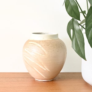 Könnte beinhalten: Eine Keramikvase mit rundem Körper und breiter Öffnung. Die Vase ist hauptsächlich hellpfirsichfarben mit einer strukturierten, gesprenkelten Oberfläche und weißen, diagonalen Pinselstrichen. Der Rand ist weiß. Eine grüne Pflanze in einem weißen Topf befindet sich im Hintergrund.