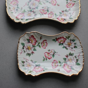 May include: Two white ceramic serving dishes with a floral pattern of pink and white roses. The dishes have a scalloped edge and a gold trim.
