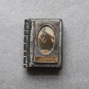 Könnte beinhalten: Ein kleines, silberfarbenes Buch mit einem Klappdeckel. Der Vorderdeckel hat einen messingfarbenen Rahmen mit einem ovalen Bild einer Person, die ein Kreuz trägt. Das Bild ist in Schwarzweiß.