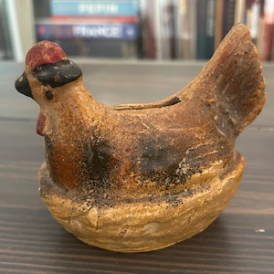 May include: A ceramic hen-shaped bank with a brown and tan finish. The hen has a red hat and black beak. The bank is sitting on a nest-like base. The bank has a slot on top for inserting coins.