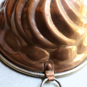 Puede incluir: Un molde para pastel de cobre vintage con un dise&ntilde;o acanalado y un lazo para colgar. El molde tiene un borde plateado.