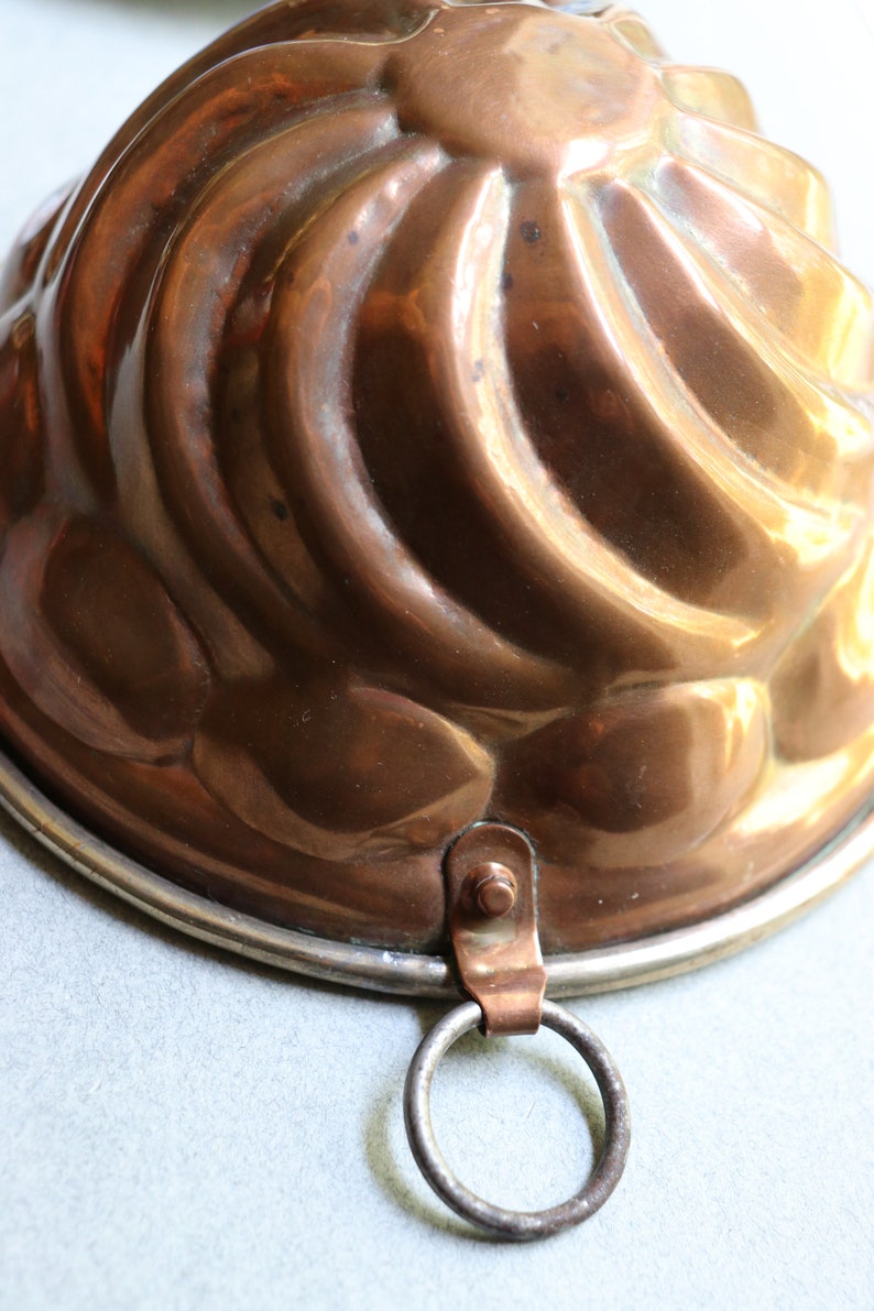Puede incluir: Un molde para pastel de cobre vintage con un dise&ntilde;o acanalado y un lazo para colgar. El molde tiene un borde plateado.