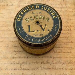Op de afbeelding: Een ronde, vintage metalen blik met een donkerblauw en goudkleurig deksel met de tekst REPRISER LUSTRE A LA PLUME L.V. PARIS 25 Grammes. De houder heeft een centraal gat in het deksel en een gestructureerde, lichtgekleurde band rond de cilindrische behuizing.